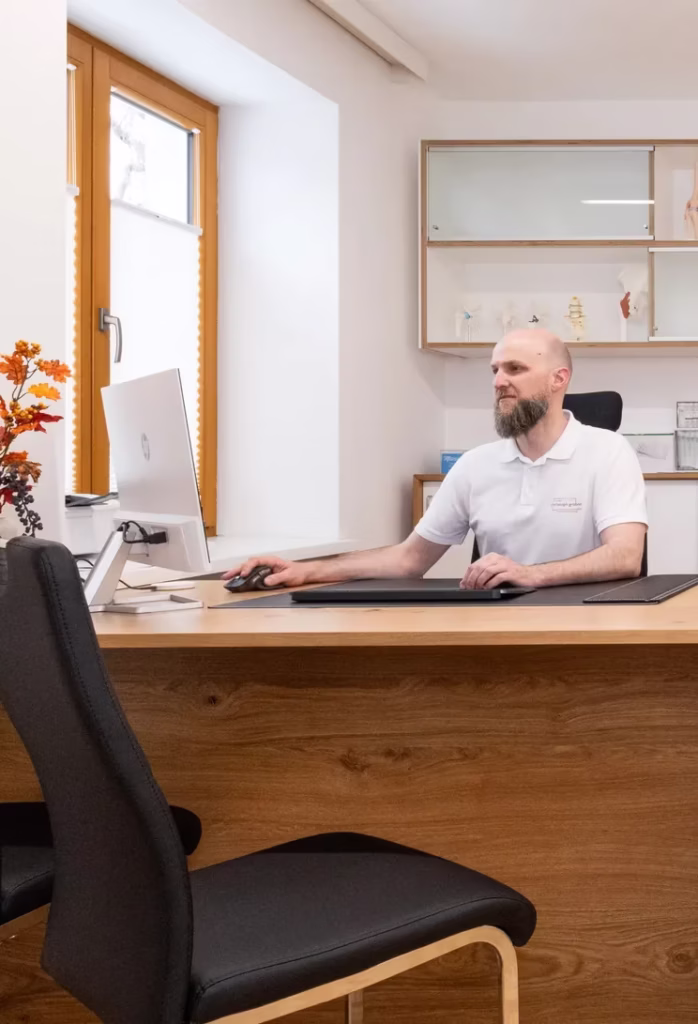 Dr Christoph Gröbner sitz an seinem Schreibtisch in seiner Praxis und schaut in den Bildschirm, er bedient dabei Maus und Tastatur